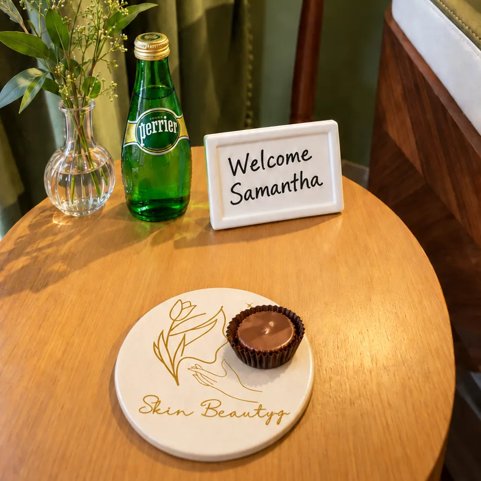 The welcome table in Suite 48: a bottle of Perrier, a small vase of greenery, a "Welcome Samantha" sign, and a dark chocolate cup on a Skin BeautyQ coaster